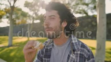 阳光明媚的一天，一个英俊的微笑的年轻人<strong>坐在公园里</strong>吃三明治-在<strong>公园里</strong>吃午饭